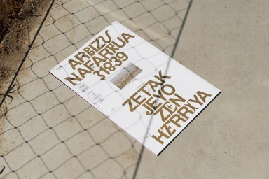 Printed poster with bold gold typography on a white paper sheet placed on a concrete surface, viewed through a wire fence.