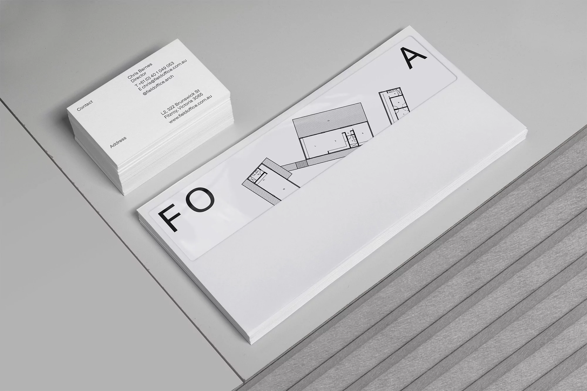 Stack of white business cards next to paper block with architectural plans and 'FOA' text, placed on a grey surface.