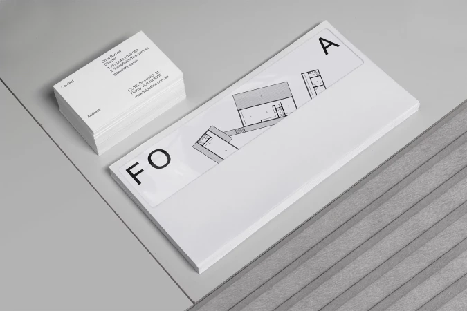 Stack of white business cards next to paper block with architectural plans and 'FOA' text, placed on a grey surface.