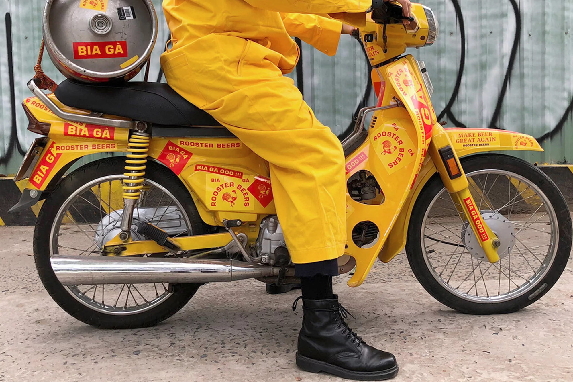 Yellow motorcycle with beer keg, 'BIA GA' stickers. Person in yellow suit riding, black boots, graffiti background. Quirky, unique vehicle design.