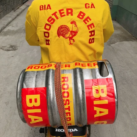 Person in yellow shirt with rooster logo carrying beer keg labeled 'Rooster Beer' and 'Bia' on back of a Honda scooter.