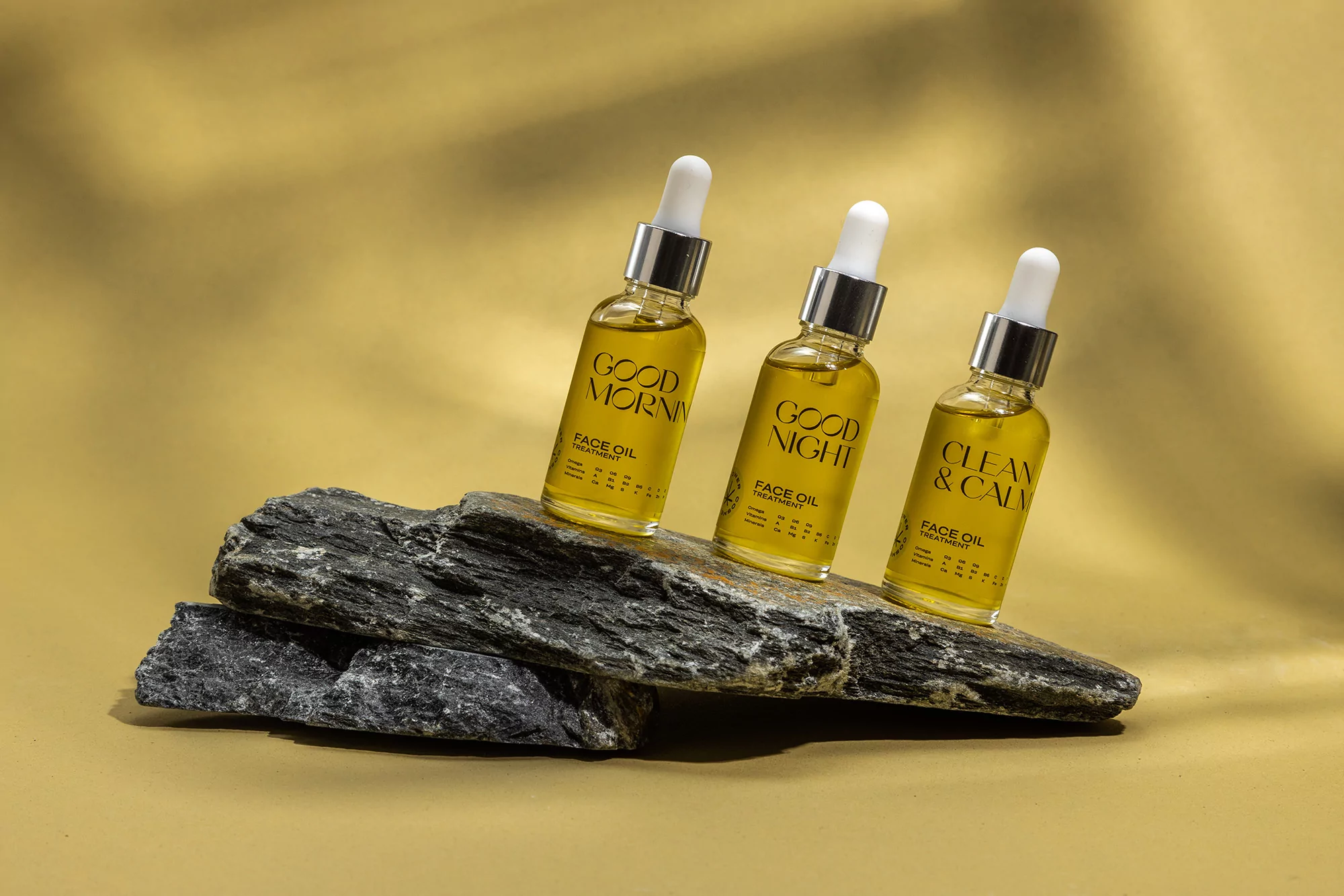 Three bottles of golden face oil with white dropper caps on a dark grey rock surface, set against a gradient gold background.
