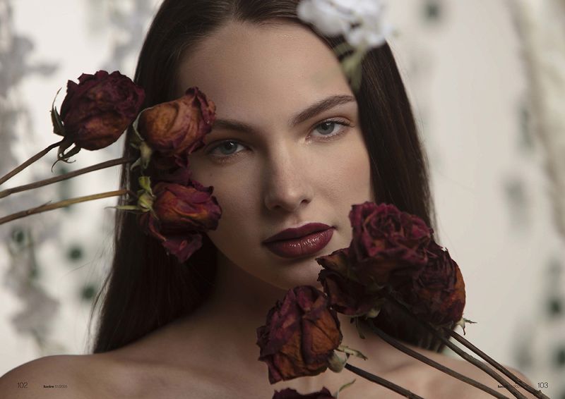 Close-up portrait of a woman with red lipstick surrounded by dried red roses, shot in a photo studio with a neutral floral background.