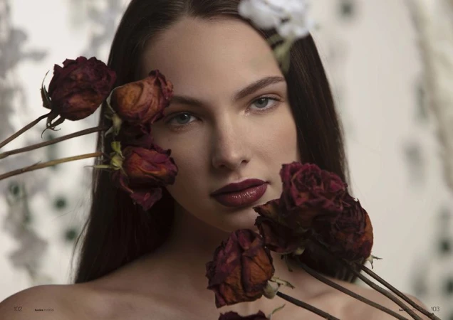 Close-up portrait of a woman with red lipstick surrounded by dried red roses, shot in a photo studio with a neutral floral background.