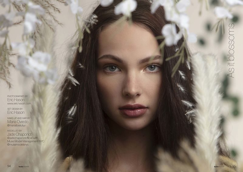 Close-up portrait of a young woman with brunette hair framed by white flowers and blurred beige foliage, under soft lighting in a studio.