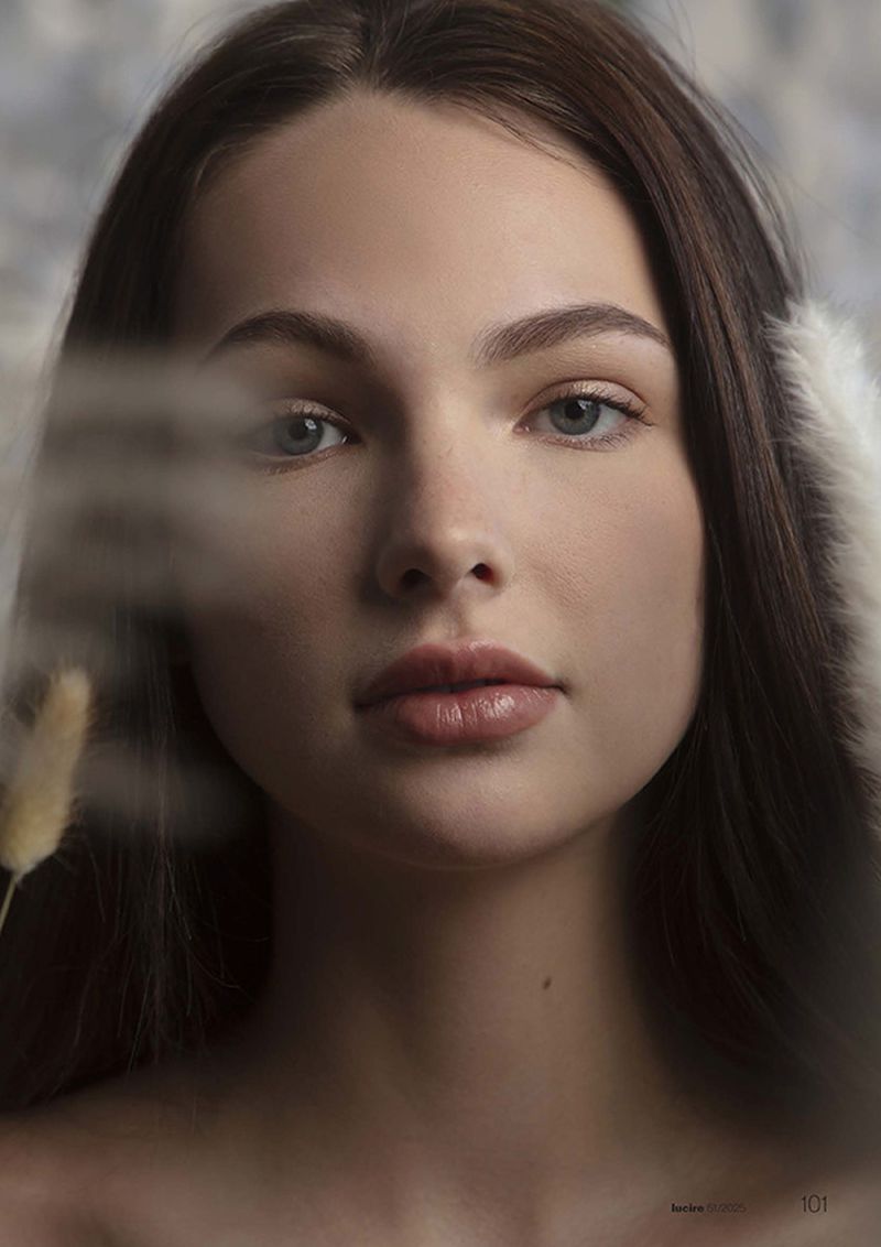 Portrait of a brunette female model with neutral expression and fair skin in soft light, complemented by a neutral-toned backdrop and feathers.