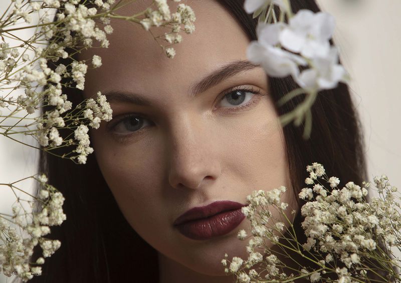 Close-up portrait of a young woman with dark hair and light eyes, framed by delicate white flowers. Burgundy lipstick completes her look.