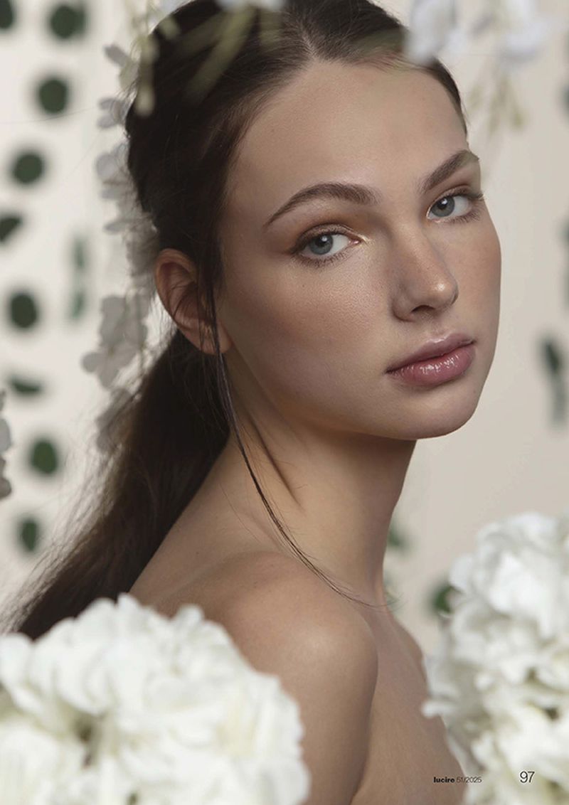 Close-up portrait of a young adult woman with light skin, light eyes, and brown hair against a floral background and soft, diffused lighting.