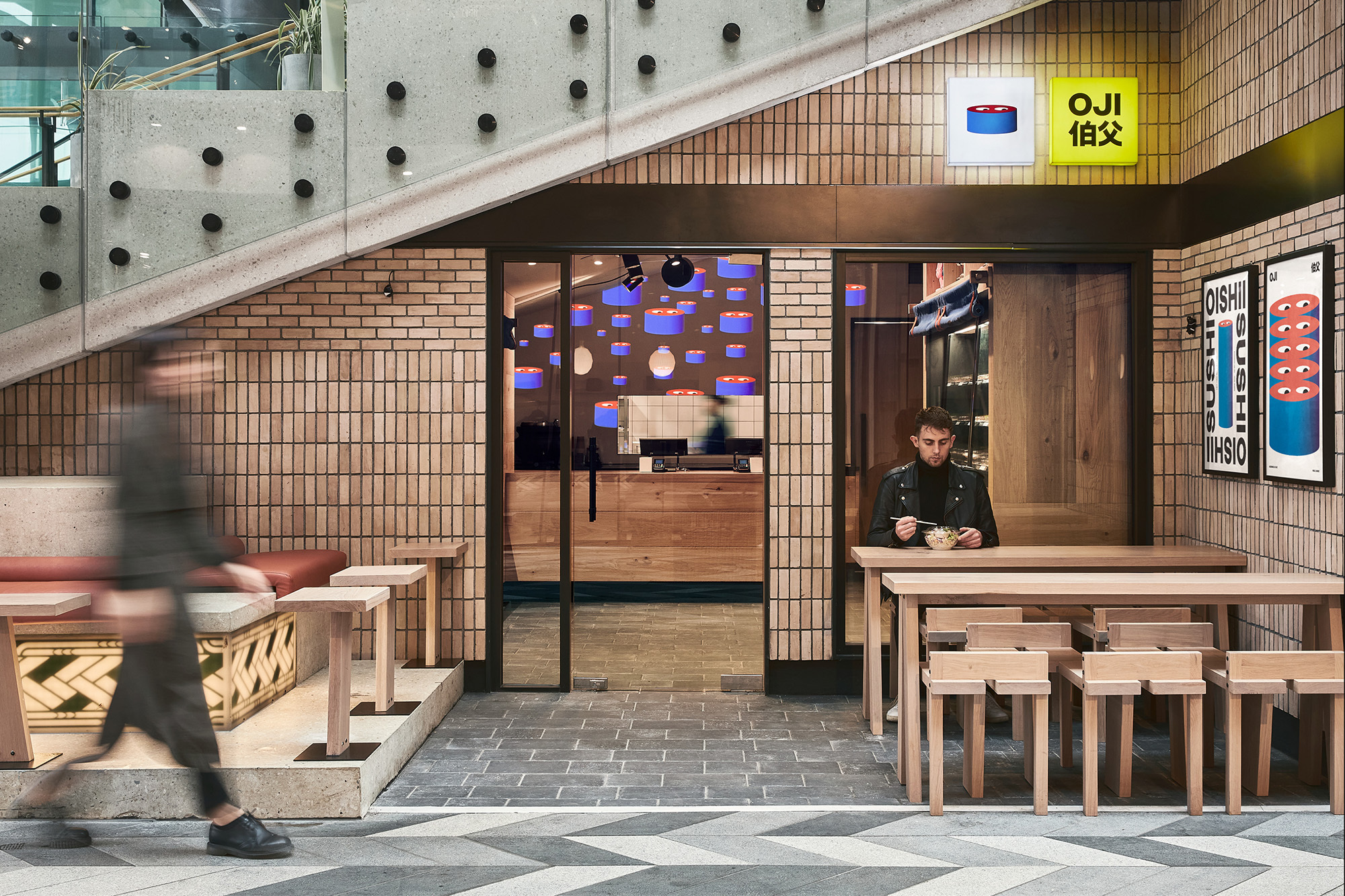 Modern sushi restaurant exterior featuring an Asian man dining, a blurred passerby, neutral brick walls, and stylish wood furniture.