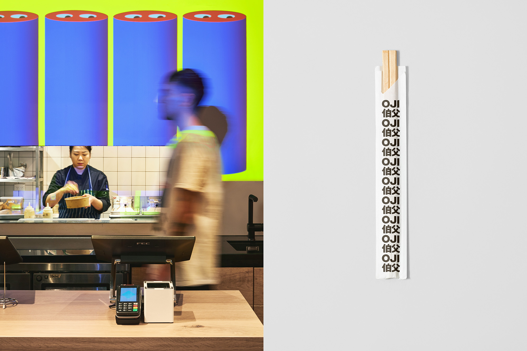 View of an open kitchen, cashier area, and white wall with chopsticks. An Asian woman prepares food, and a blurred person walks by.