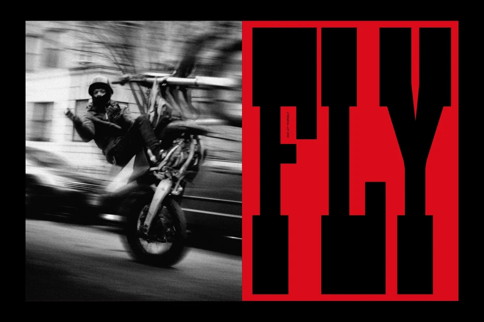 Monochrome image of a man doing a wheelie on a motorcycle on an urban street next to a large, bold red and black graphic design.