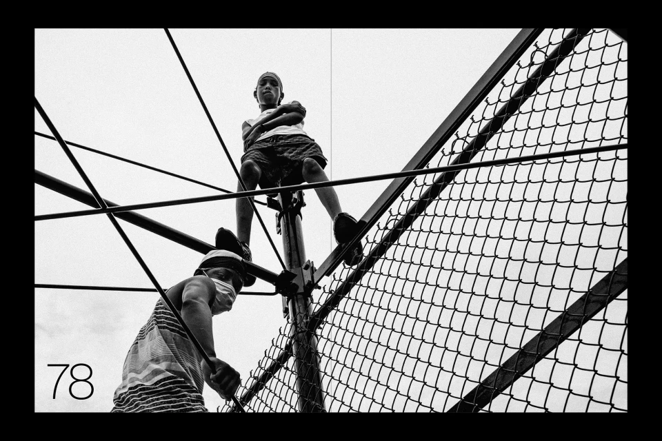 Low angle, black and white shot of two children climbing on a playground structure with a chain-link fence in the background.