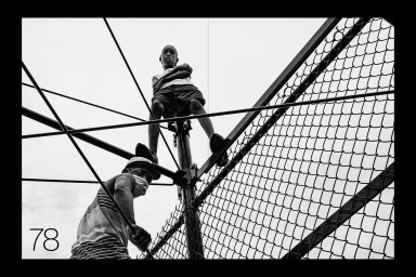 Low angle black and white photograph of two young males climbing a chain-link fence structure under a bright overcast sky.