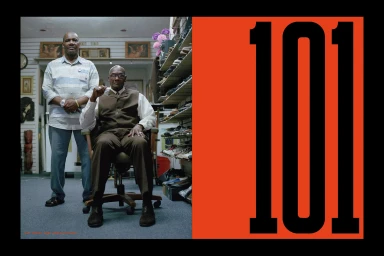Two Black men in a retail store interior alongside a large orange panel displaying the bold black number 101.