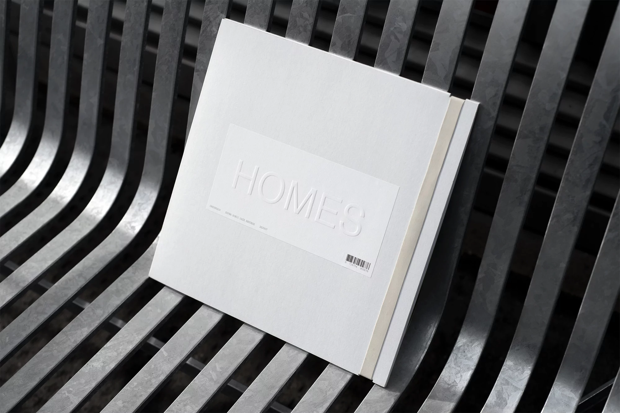 A stack of white books titled 'Homes' is placed on a modern metal bench, with a focus on simple, clean design and monochromatic color scheme.