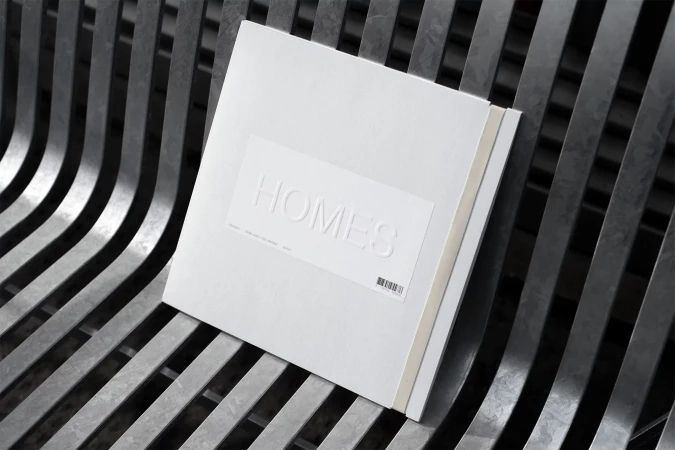 A stack of white books titled 'Homes' is placed on a modern metal bench, with a focus on simple, clean design and monochromatic color scheme.