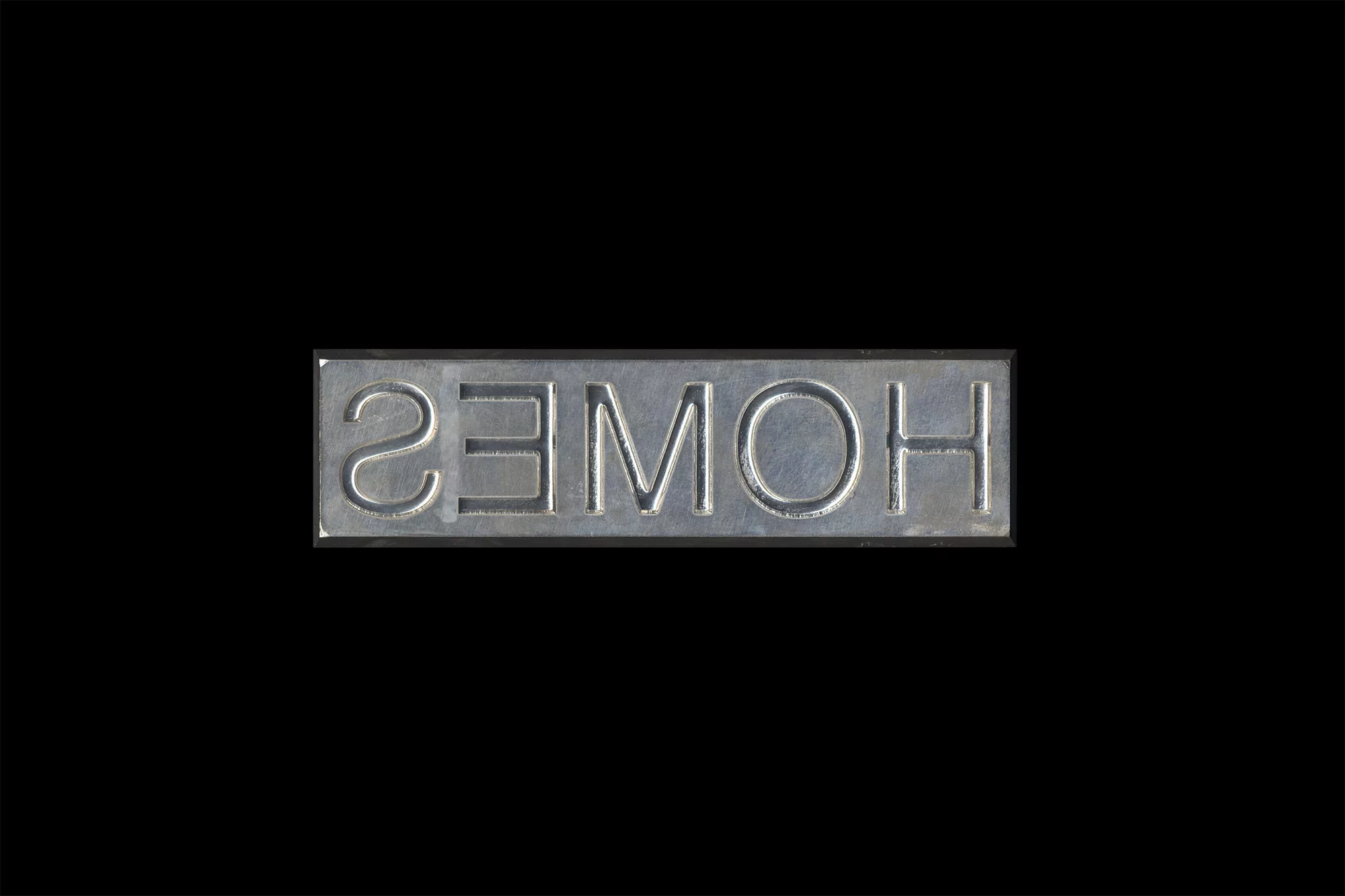Metal sign with the word 'HOMES' embossed in capital letters against a black background in a studio product shot.