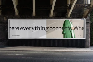 Large outdoor billboard advertising display mounted under a concrete bridge showing typography and a green coat