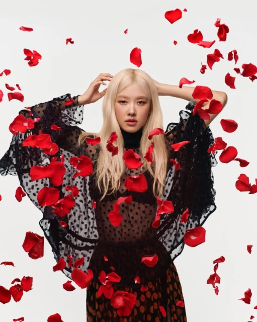 Portrait of a blond adult woman wearing a black polka-dot dress, posing with her hands in her hair, surrounded by red rose petals.