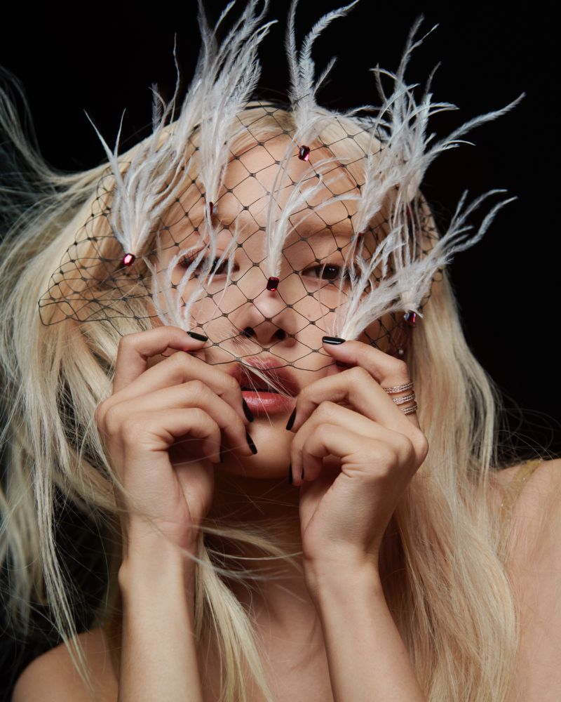 Woman with blonde hair and feather headpiece holds netting in a portrait against a dark backdrop, showcasing beauty and fashion.