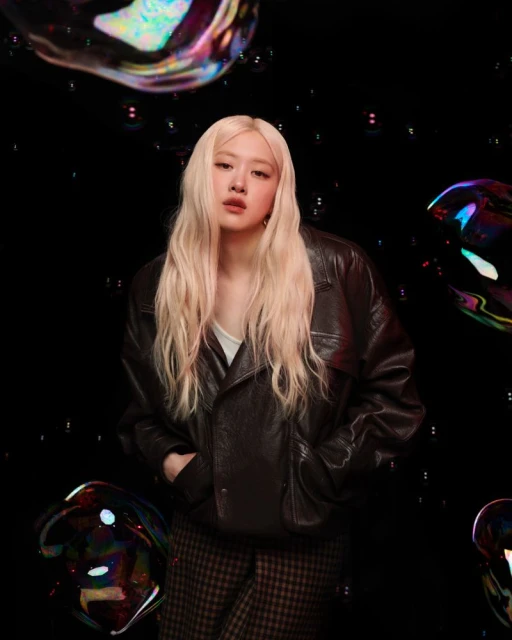 Blonde haired woman with jacket posing in front of black background with transparent iridescent bubble objects floating throughout the scene.
