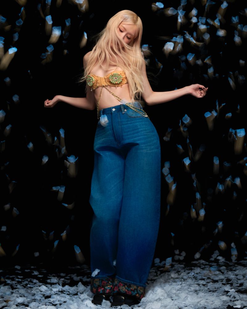 Young woman with long blonde hair wearing jeans and a gold top poses in a dark studio with out-of-focus light effects.
