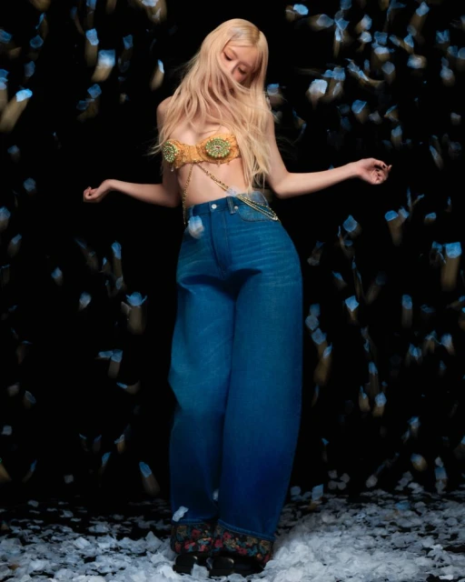 Young woman with long blonde hair wearing jeans and a gold top poses in a dark studio with out-of-focus light effects.