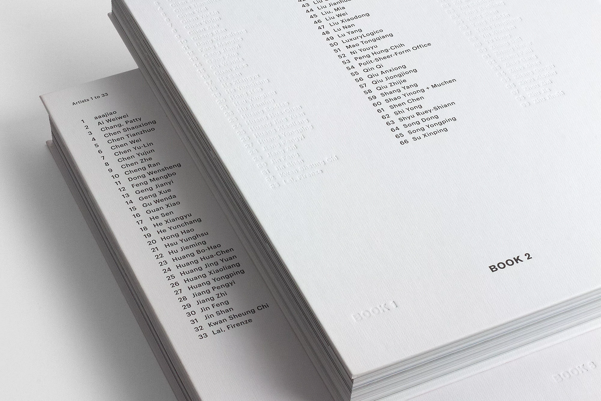 Two open books display lists of artist names and numbers in a white on white aesthetic, showing minimalistic design and information.