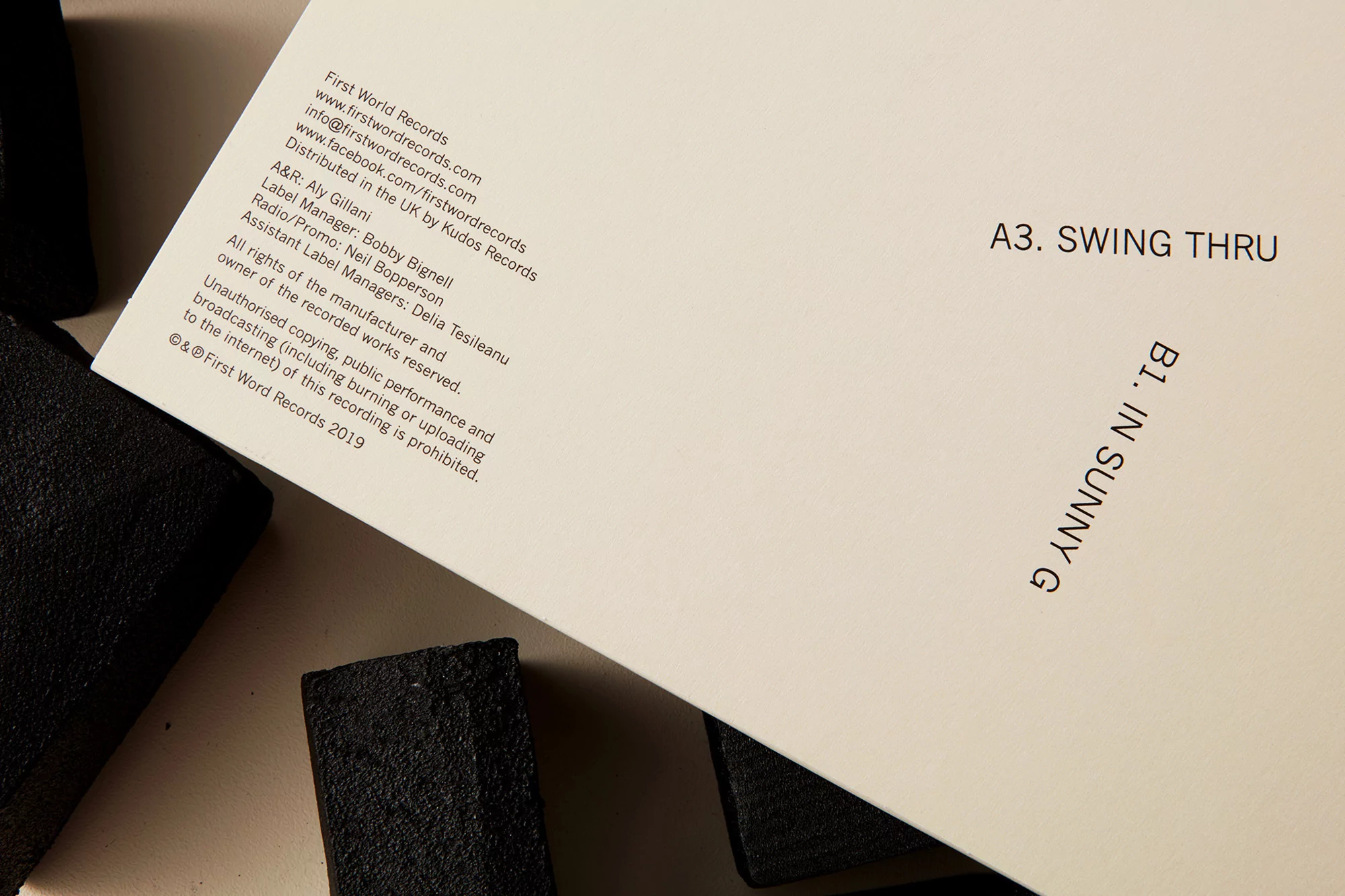 Vinyl record sleeve featuring track titles and record label information, set against a plain background with textured black objects.