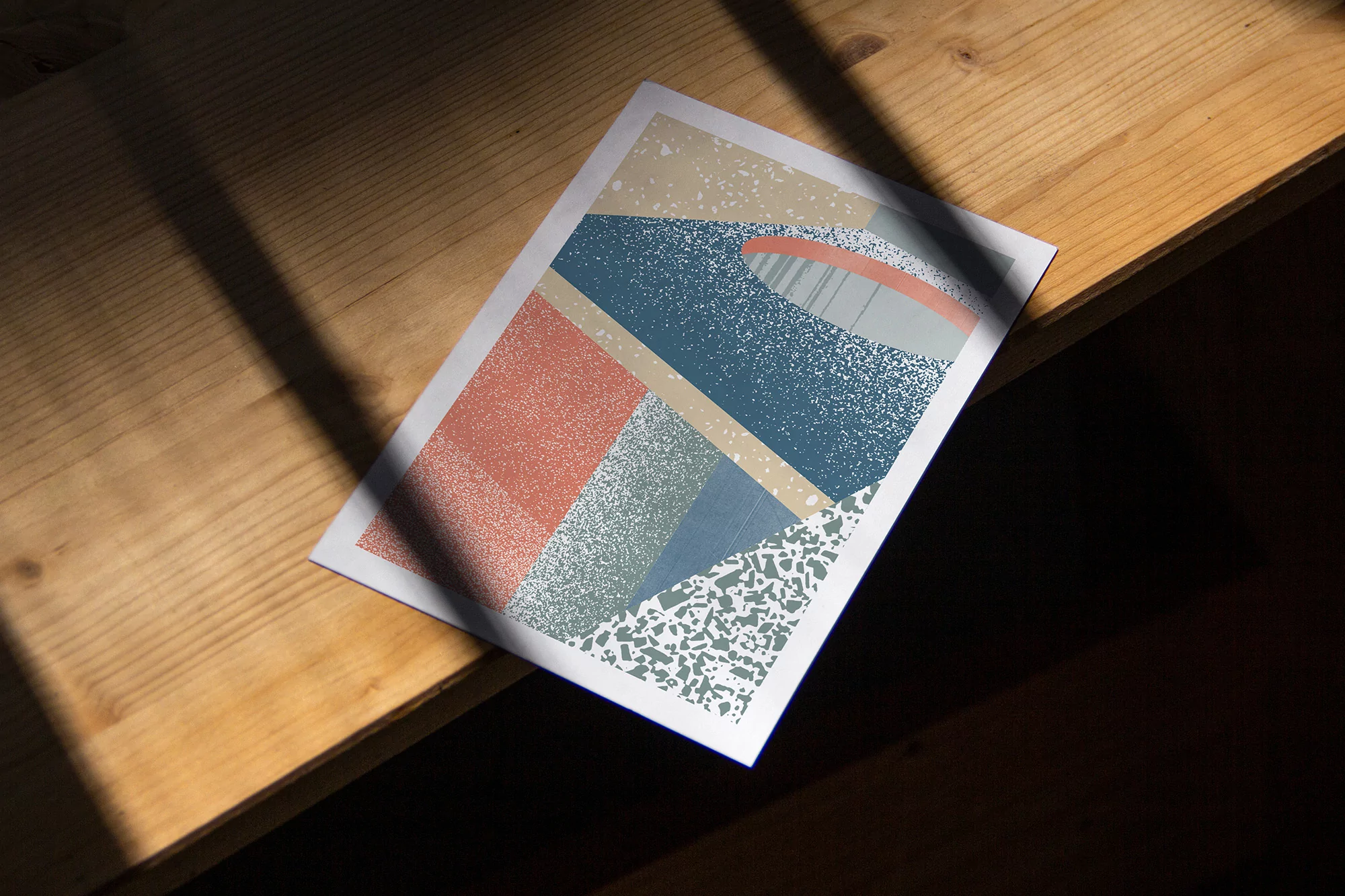 Abstract geometric art print in shades of blue, tan, orange, and green on a light wood table with natural sunlight and shadows.