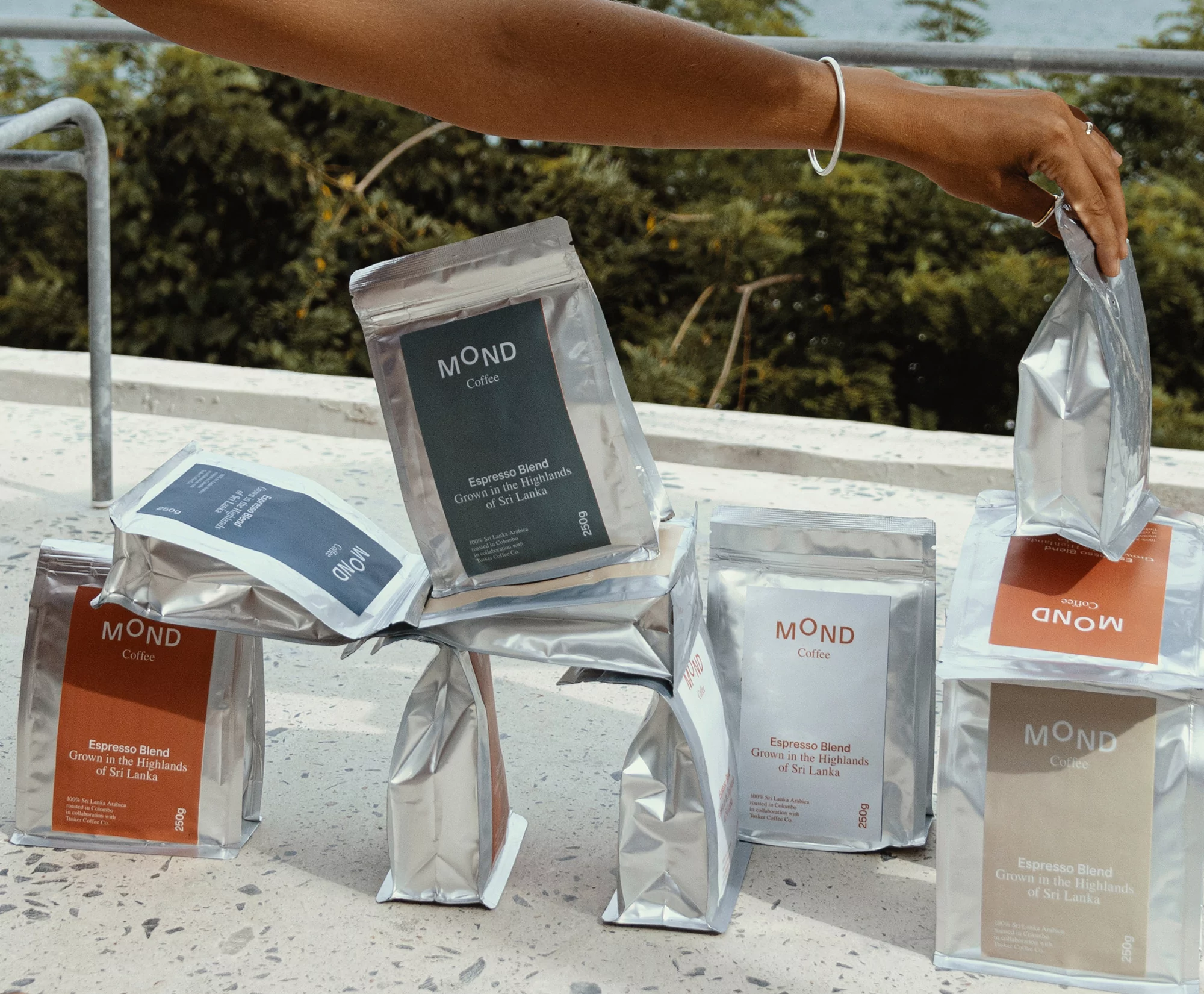 Hand arranges Mond Coffee packages outdoors. Silver foil bags with dark and tan labels stacked on a concrete surface with a green background.