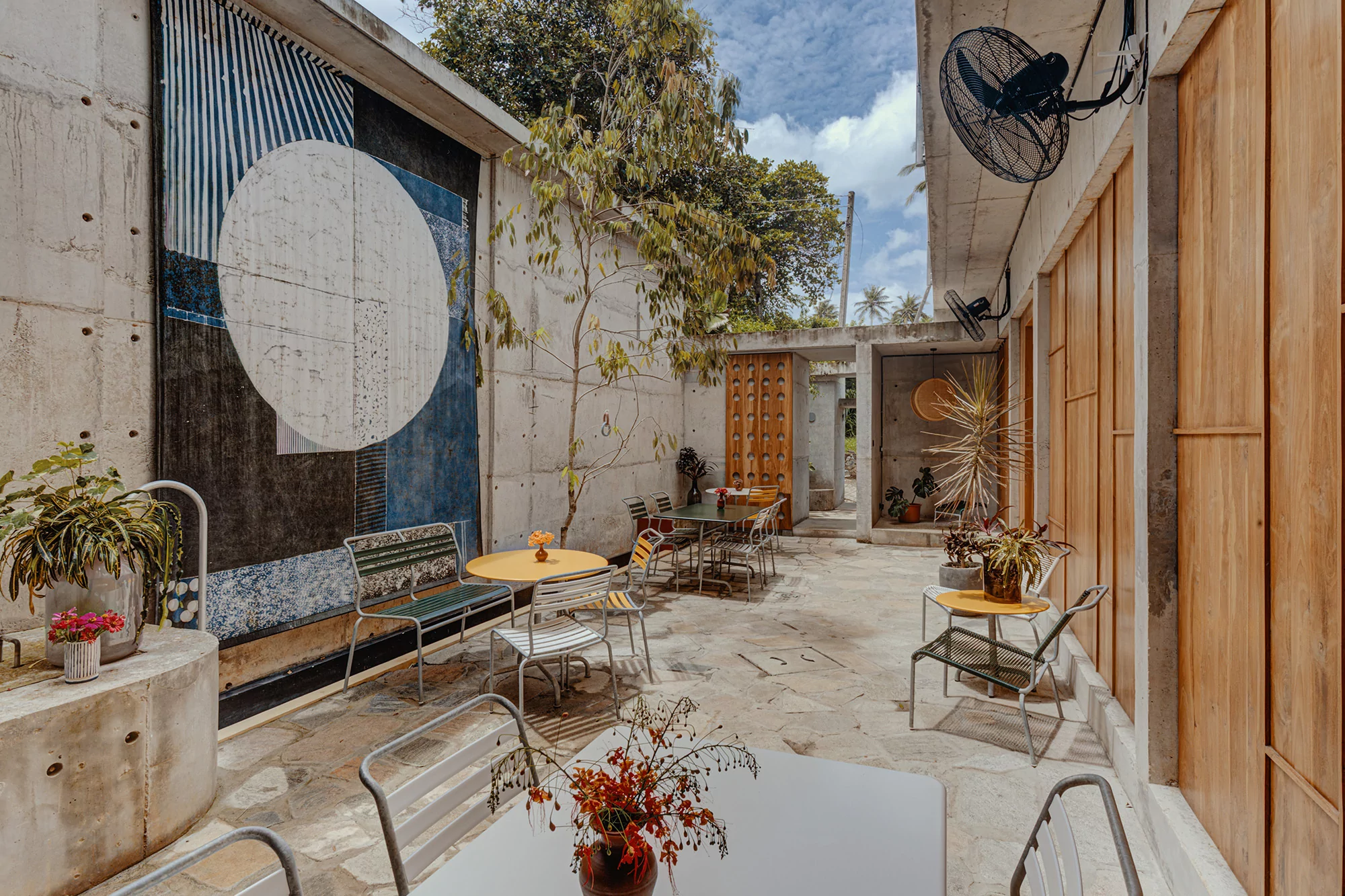 Outdoor patio with architectural concrete walls, tables and chairs, and abstract art, featuring plants and trees under a bright sky.