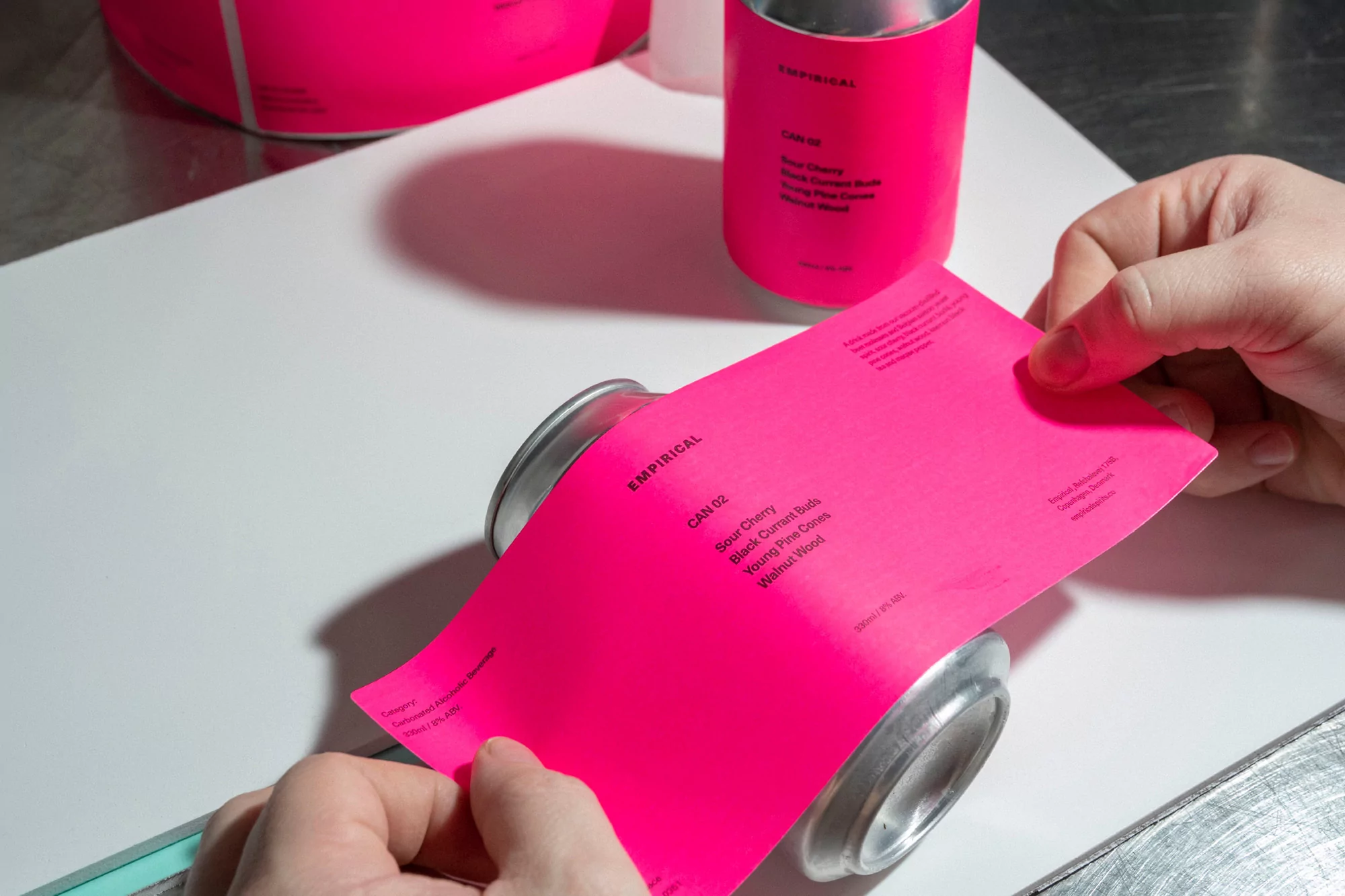 A person affixes a bright pink label onto a silver beverage can, featuring text elements; additional cans and labels surround them.