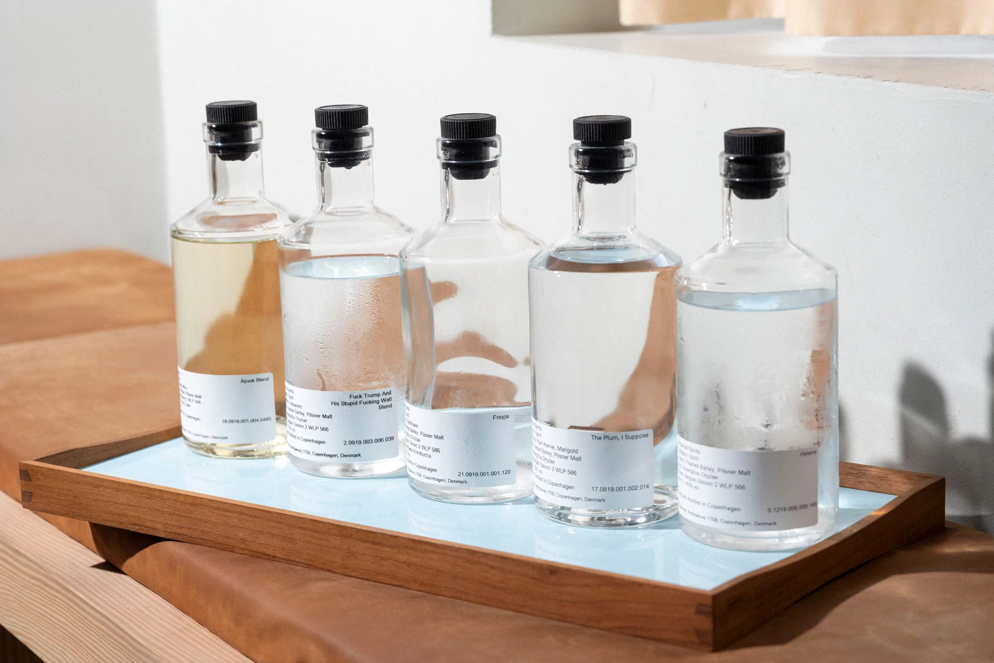 Five clear glass bottles with black stoppers filled with liquids on a light blue tray sitting on a tan upholstered surface.