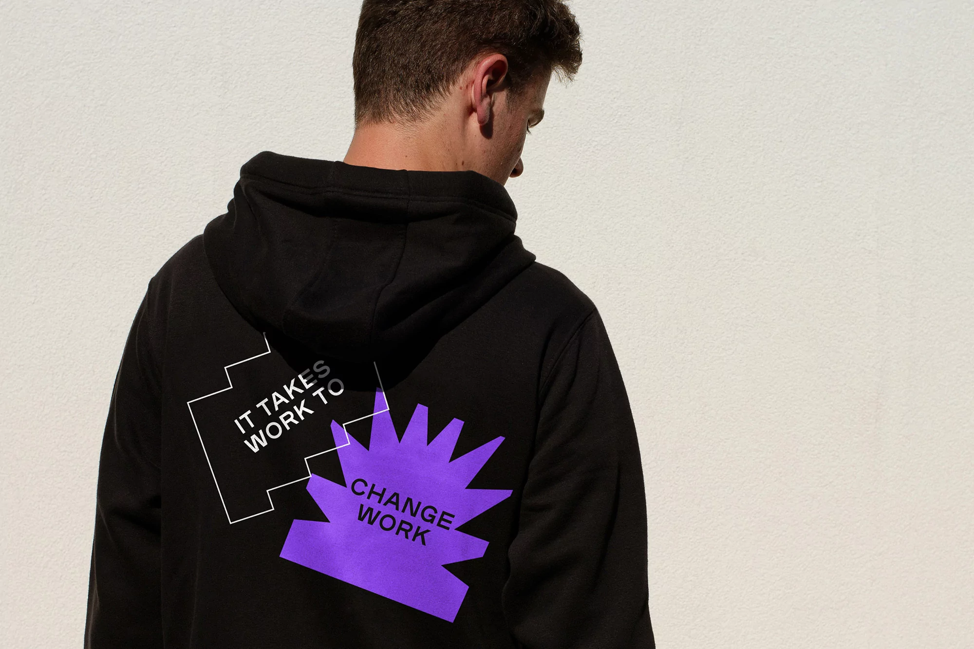 Back view of a young adult wearing a black hooded sweatshirt with purple and white graphic text on the back against a white background.