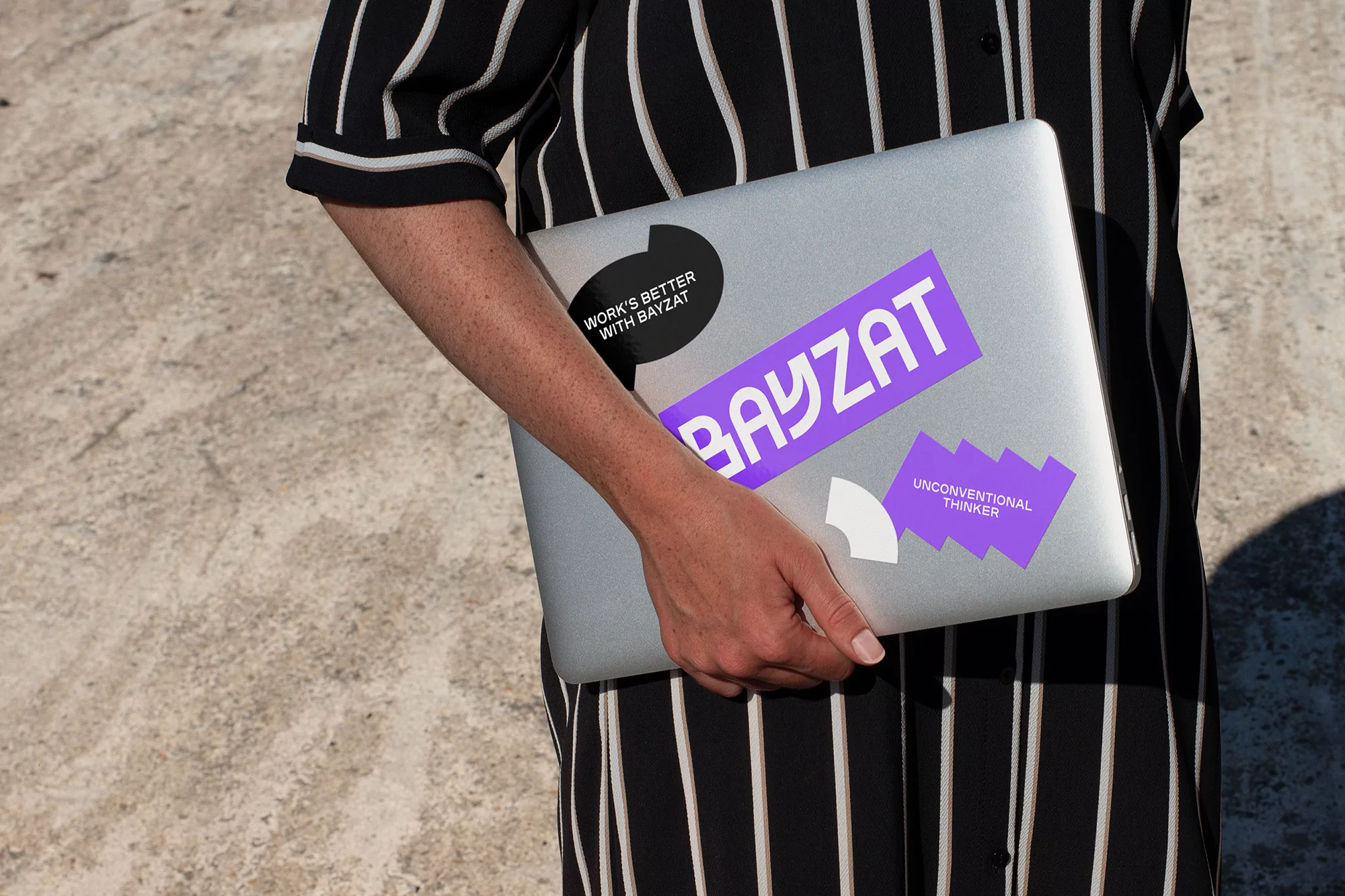 Person holds an open silver laptop adorned with stickers in front of a vertically striped black and white shirt against a concrete background.