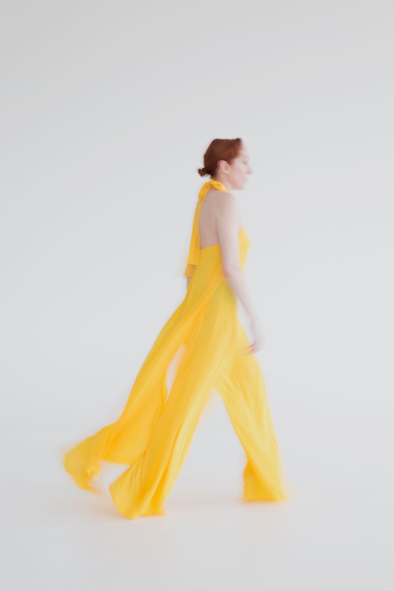 Woman walks in flowing yellow dress in a minimalist studio. The garment and motion blur create a dynamic feel against the white backdrop.