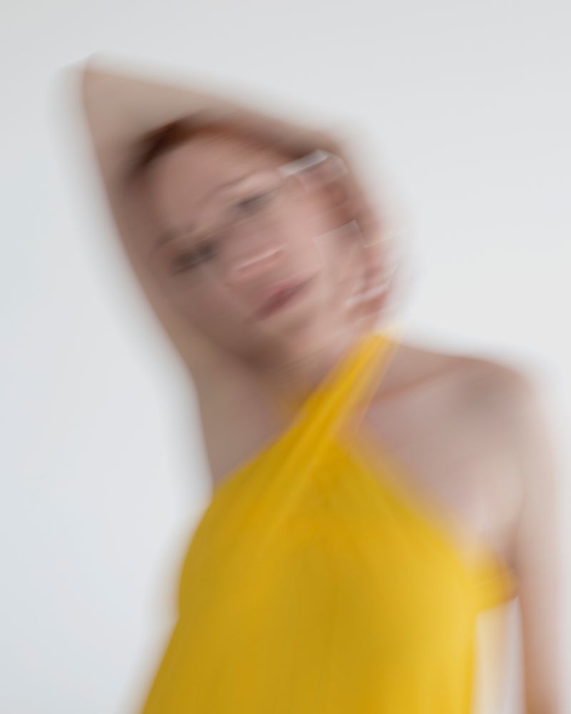 Blurred portrait of a woman in yellow top against a white background with soft lighting and intentional motion blur.