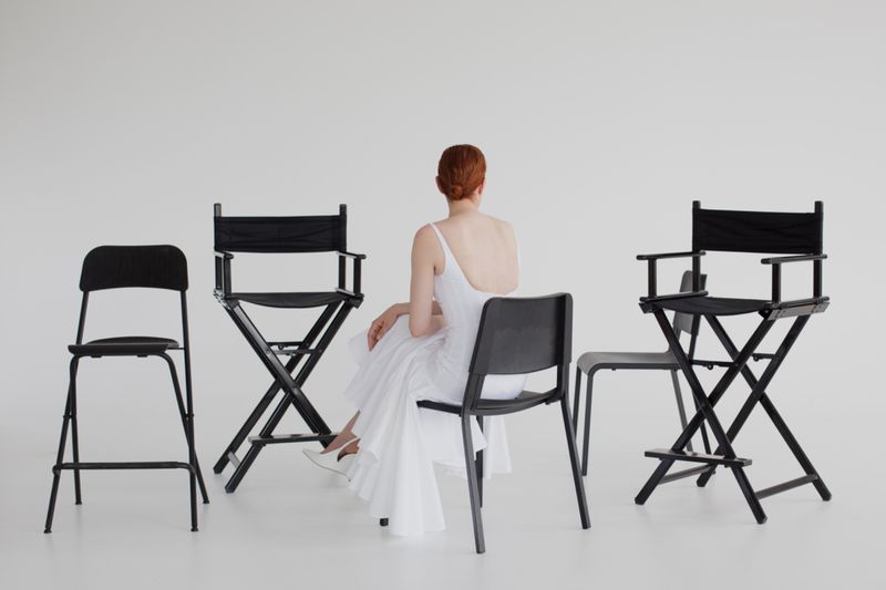 Back view of a woman with red hair in a white dress seated on a black chair surrounded by director's chairs in a white room.