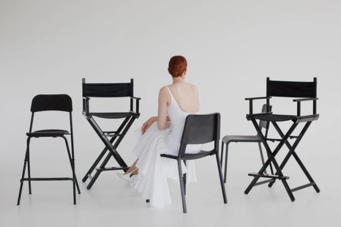 Back view of a woman with red hair in a white dress seated on a black chair surrounded by director's chairs in a white room.