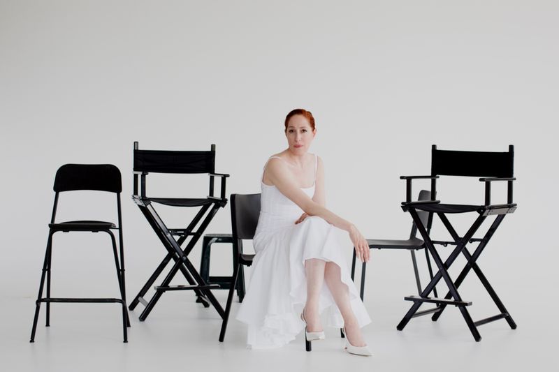 A woman with red hair sits on a chair, wearing a white dress, posed in a studio setting with black director's chairs surrounding her.