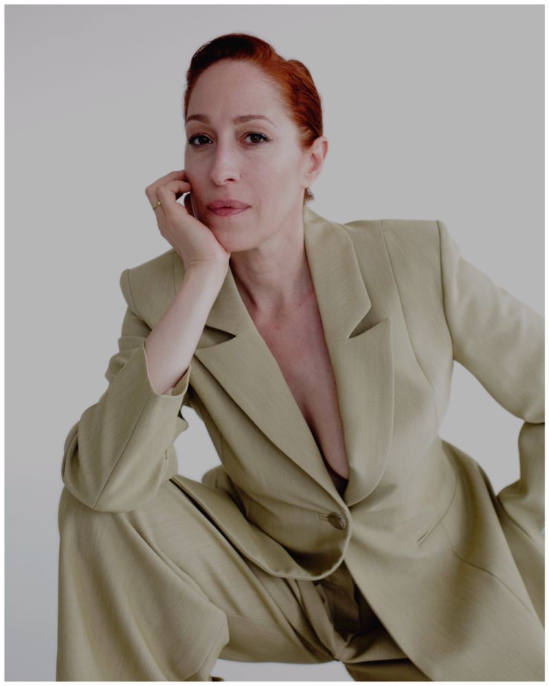 Adult woman with red hair poses in a tan suit in a studio. Portrait showcases fashion and beauty with a minimalist studio setting.