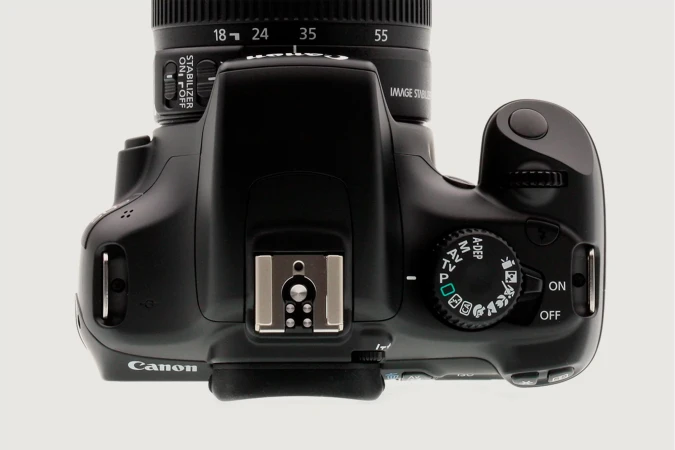 Overhead shot of a black Canon DSLR camera showcasing its lens, settings dial, hot shoe, and various controls on a white background.