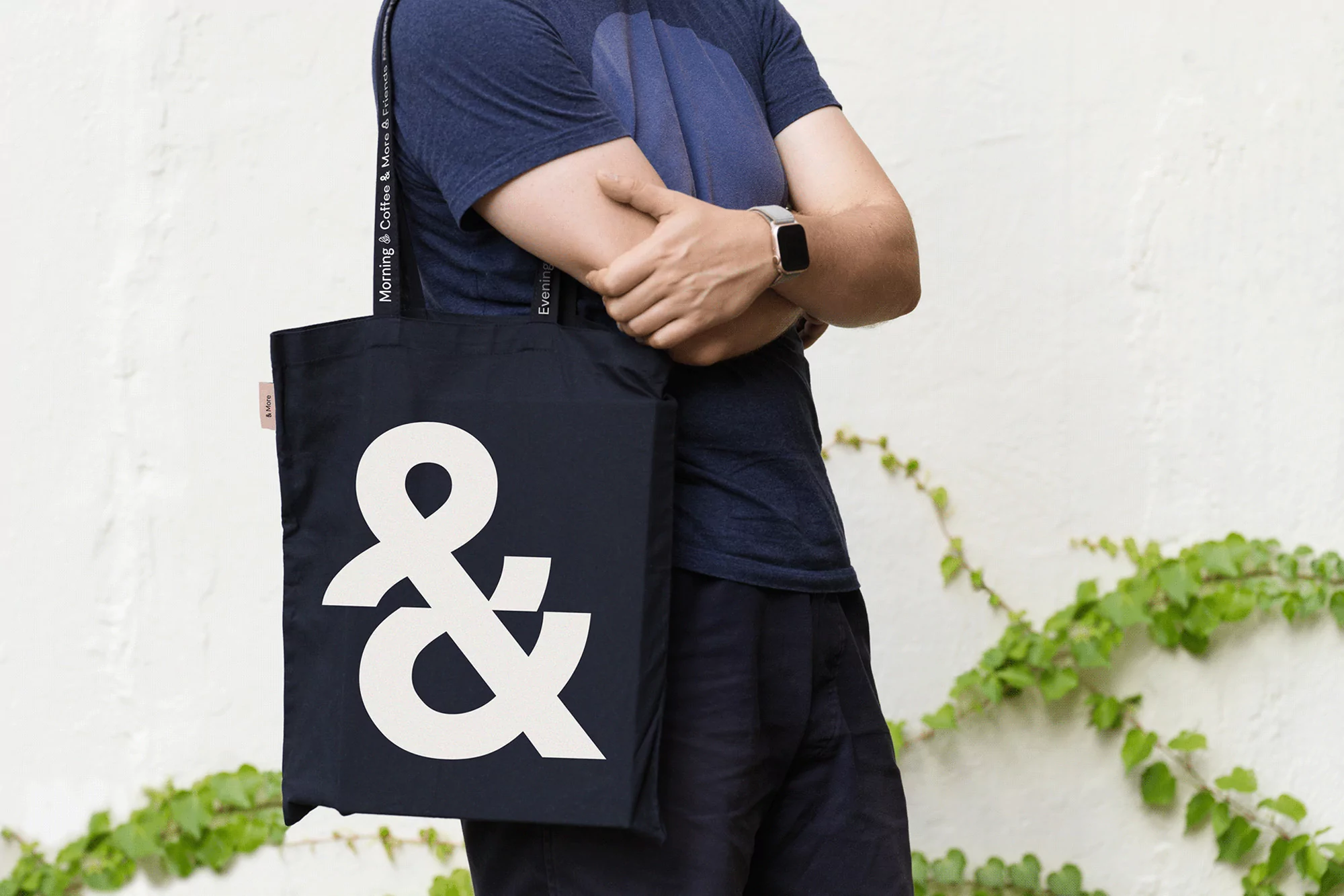 Person with a navy tote bag featuring a large white ampersand, against a textured white wall with green vines. Stylish and functional.