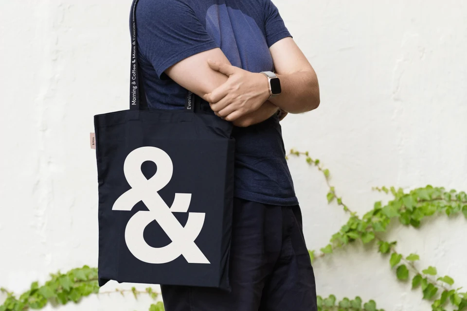 Person with a navy tote bag featuring a large white ampersand, against a textured white wall with green vines. Stylish and functional.