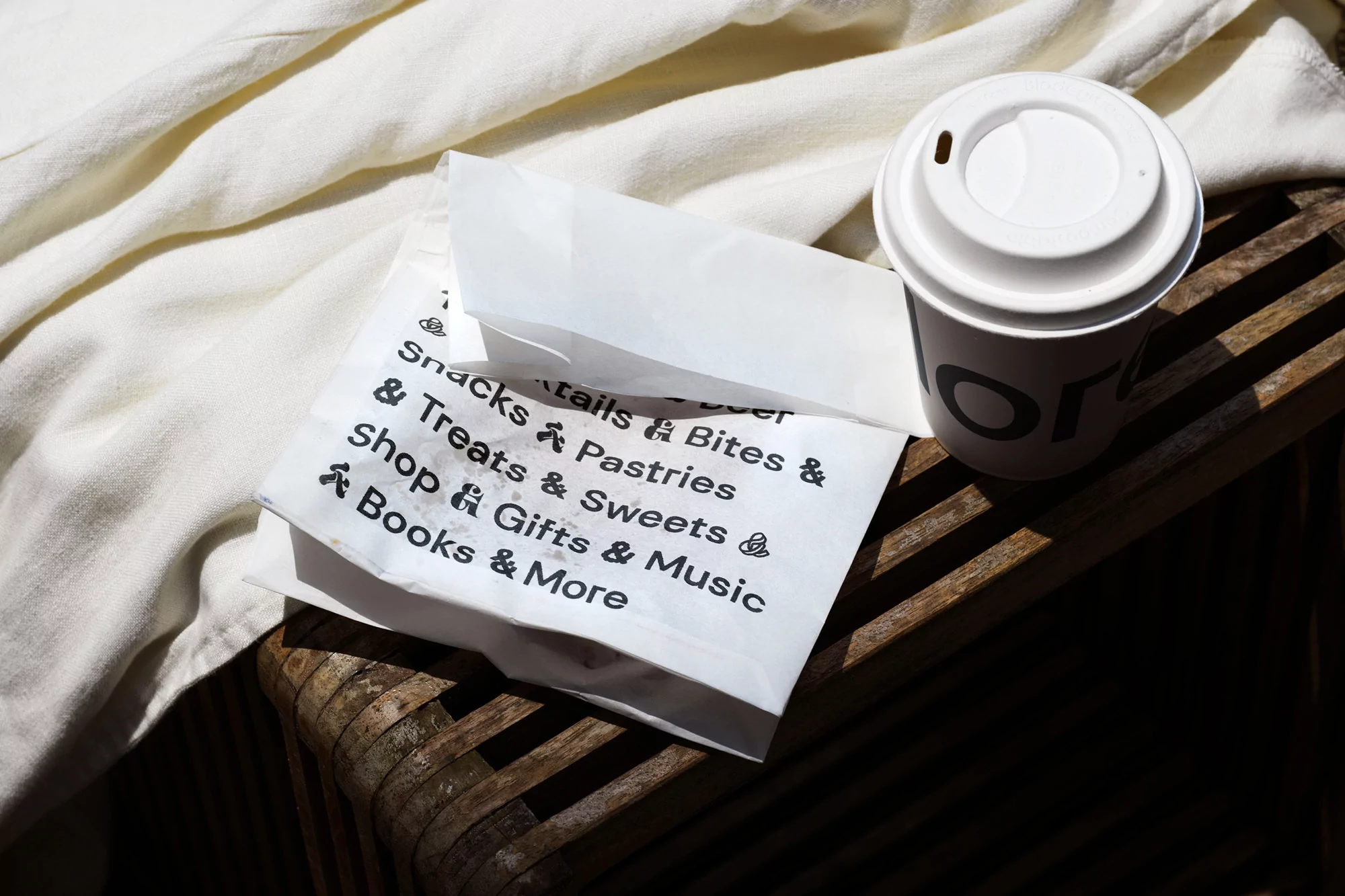 Overhead shot of a white takeout coffee cup, a paper bag with food shop text, all resting on a wooden stool with a cream-colored cloth.