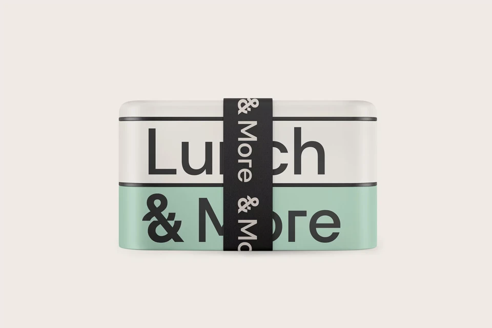 Lunch box with mint green and white layers, secured by black band with '& More' text, sitting on a light beige background.