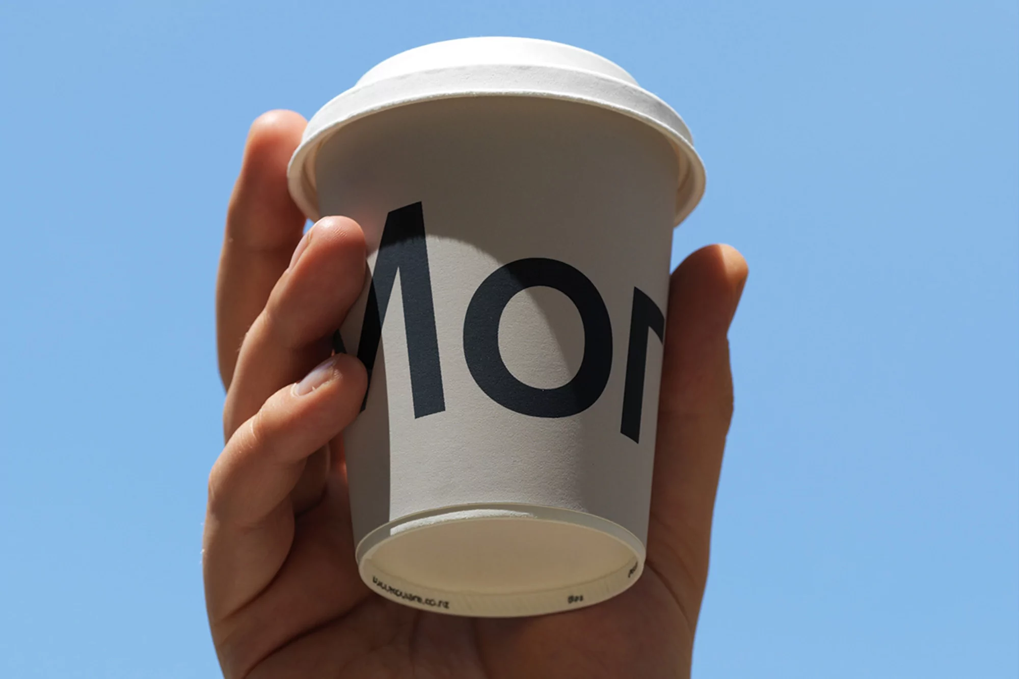 Close-up of a hand holding a white disposable coffee cup against a bright blue sky, showcasing the cup's minimalist design and brand logo.