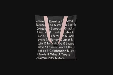 Black tote bag featuring white printed typographic words and pink shoulder straps on dark background