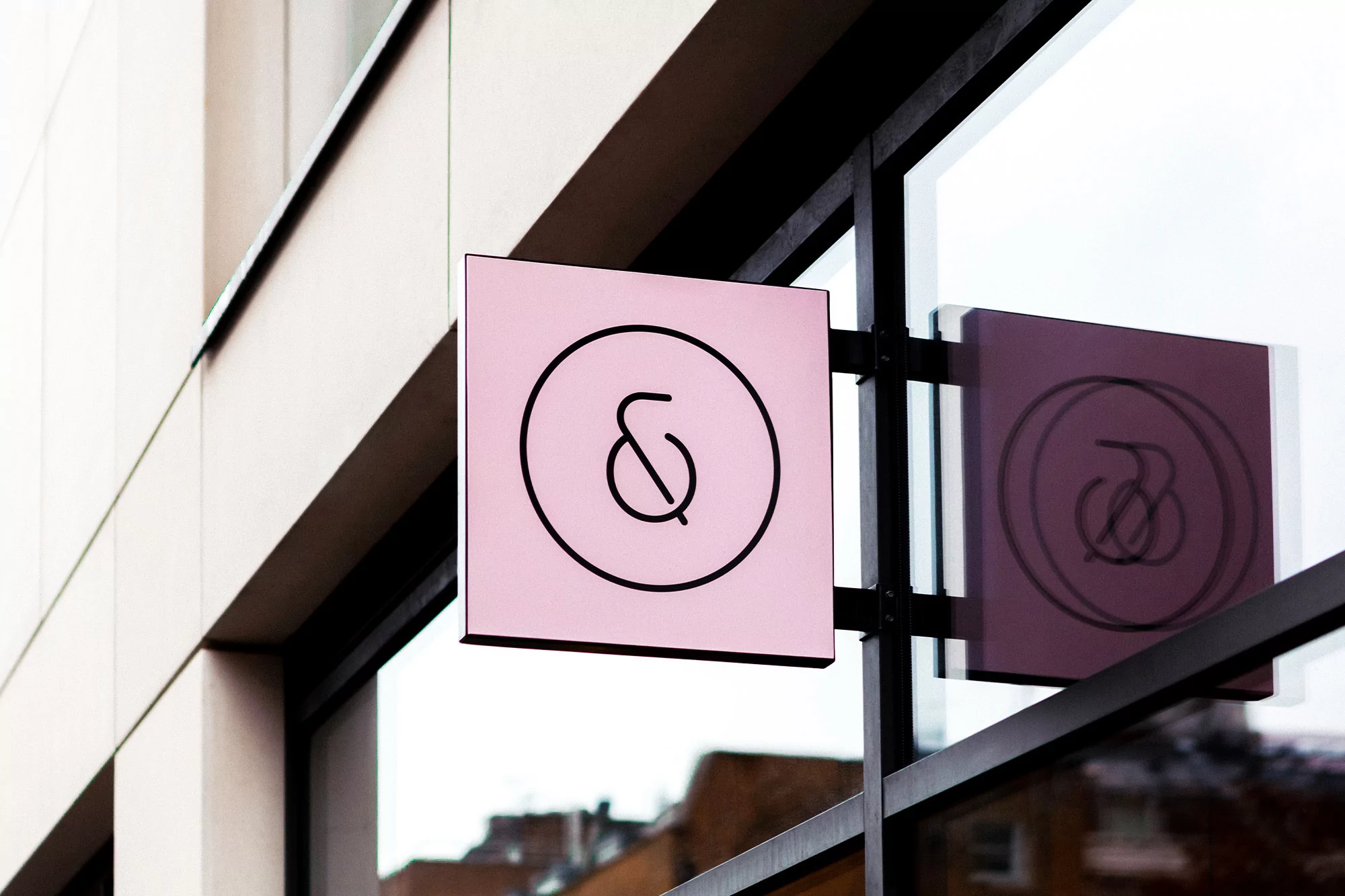 A pink square sign featuring a stylized ampersand hangs outside a building with glass windows, reflecting the sign itself and the surroundings.